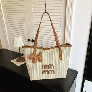 A woven straw tote bag with tan leather handles and a lettered charm, standing on a dark box